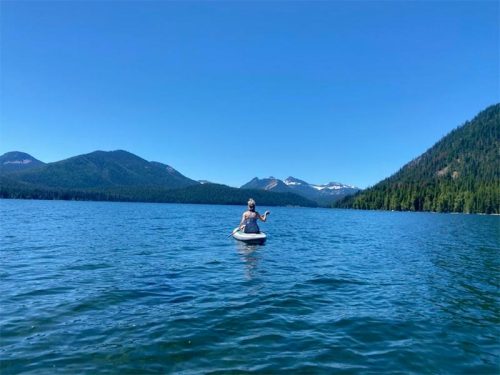A person in a small boat paddles on a calm lake surrounded by mountains and clear blue skies.
