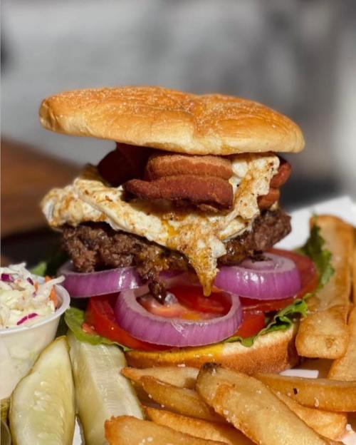 A hearty burger with a fried egg, crispy bacon, lettuce, tomato, and onion, served with fries and pickles on the side.