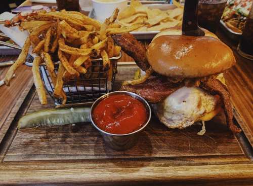 A burger with bacon and a fried egg, served with fries in a basket, a pickle, and a small cup of ketchup on a wooden board.