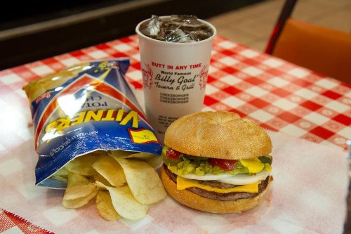 A cheeseburger with toppings, a bag of potato chips, and a cup of soda on a checkered tablecloth.
