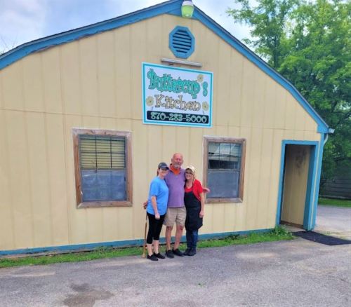 Three people stand in front of a yellow building with a sign that reads "Buttercup's Kitchen."