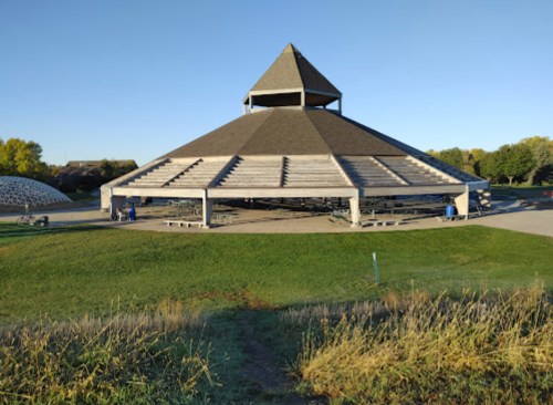 A large, pyramid-shaped building with a sloped roof, surrounded by green grass and trees under a clear blue sky.