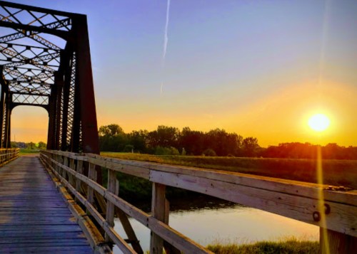 A sunset view from a wooden bridge, with a river and trees in the background under a clear sky.