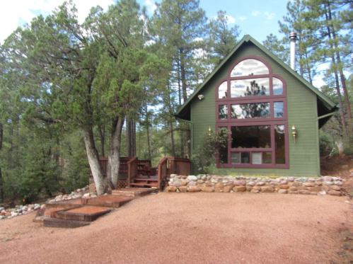 A green cabin with large windows surrounded by trees and a gravel path, featuring a wooden deck and stone landscaping.