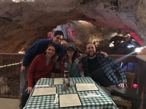 A group of four friends poses at a table in a cave restaurant, with rock formations and colorful lights in the background.
