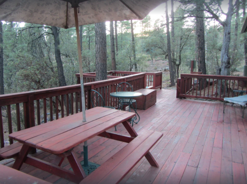 A wooden deck surrounded by trees, featuring a picnic table, a small table, and an umbrella.