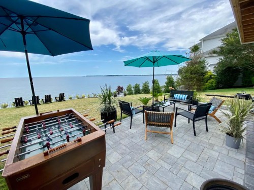 A lakeside patio with a foosball table, seating areas, and umbrellas, surrounded by greenery and water views.