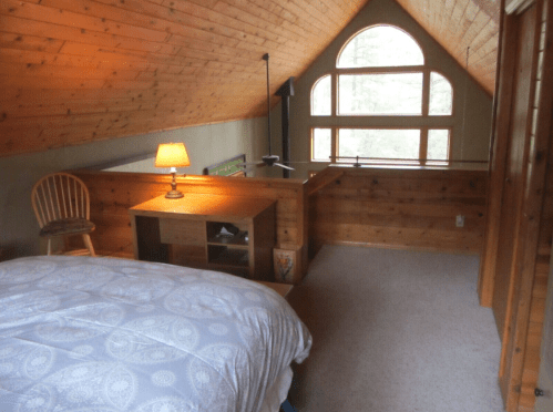 Cozy attic bedroom with a wooden ceiling, a bed, desk, chair, and a large window letting in natural light.