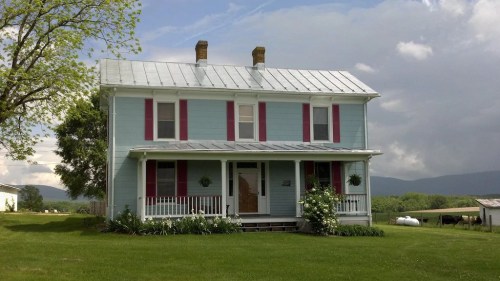 A light blue two-story house with red shutters, a metal roof, and a green lawn, set against a cloudy sky.