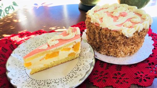 A round cake with pink frosting and a slice on a plate, showcasing layers of cream and fruit. Decorated with swirls.