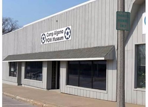 Exterior of the Camp Algona POW Museum, featuring a sign and large windows, set on a paved street.
