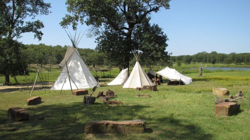 Three white tipis set up in a grassy area near a pond, surrounded by stones and trees.
