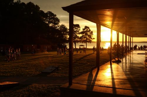 Silhouetted figures gather near a lakeside pavilion at sunset, with warm light casting long shadows on the ground.