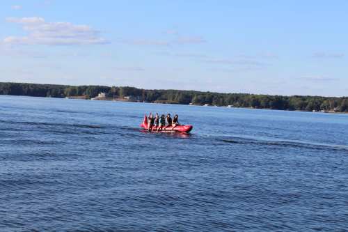 A group of six people in a red boat on a calm lake, surrounded by trees and blue sky.