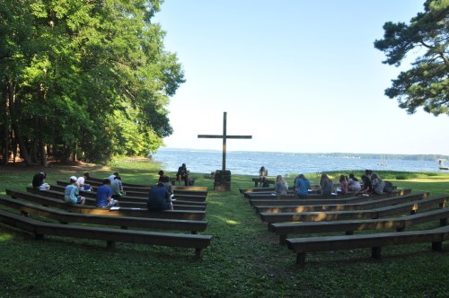 A group of people sitting on benches in a circle around a cross by a lake, surrounded by trees.