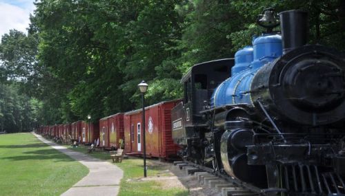 A vintage steam locomotive beside red train cars along a tree-lined path in a park setting.