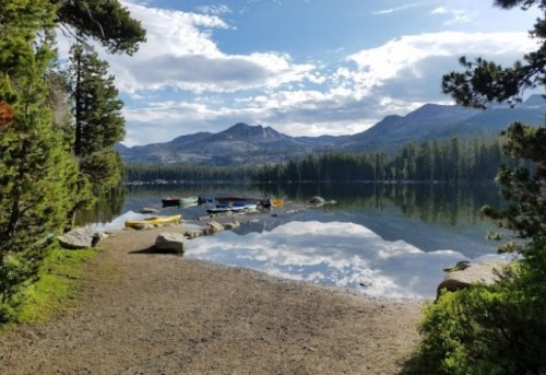 Serene lake scene with colorful kayaks, surrounded by trees and mountains, reflecting in calm water under a cloudy sky.