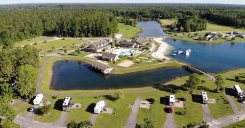 Aerial view of a campground with a pool, sandy beach, and surrounding greenery, featuring RVs parked along the pathways.
