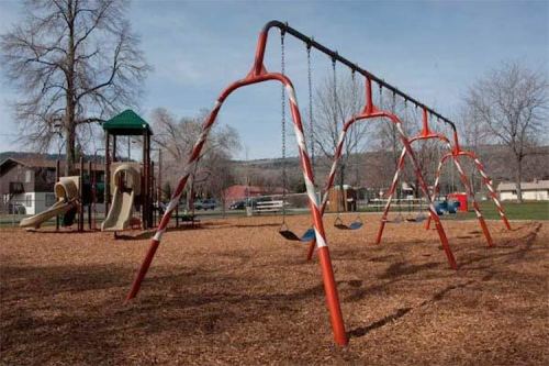 A playground featuring swings and a slide, surrounded by wood chips and trees under a clear sky.