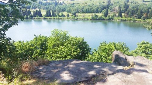 A serene view of a calm river surrounded by lush green trees and rocky terrain under a clear blue sky.