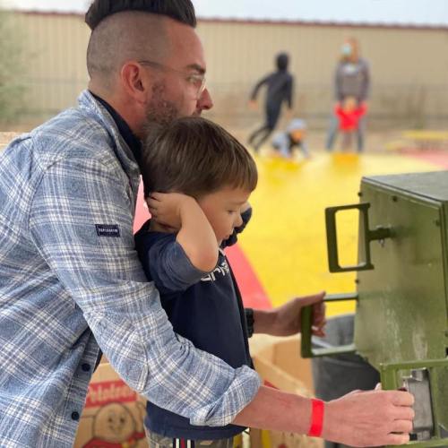 A man and a young boy interact at a playground, with children playing in the background. The man is wearing a plaid shirt.