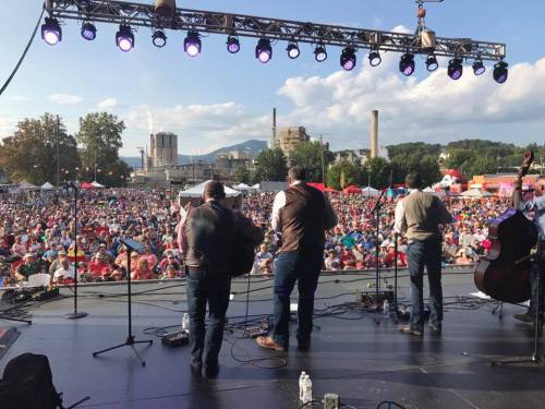 A crowd enjoys a live music performance outdoors, with musicians on stage and a scenic backdrop of mountains and buildings.