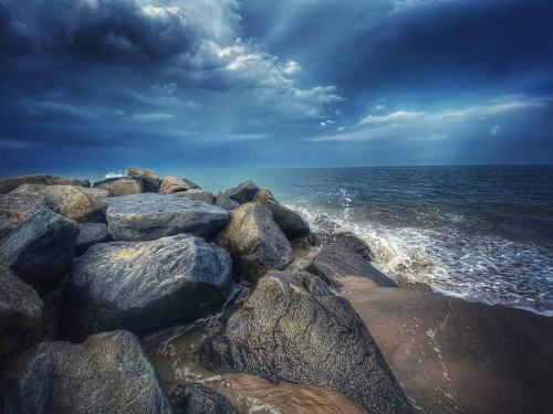 Rocky shoreline with waves crashing against the stones under a dramatic, cloudy sky.