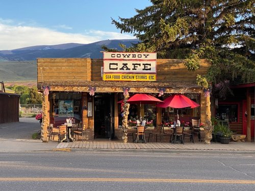 A rustic café with a wooden exterior, red umbrellas, and mountain scenery in the background. Sign reads "Cowboy Café."