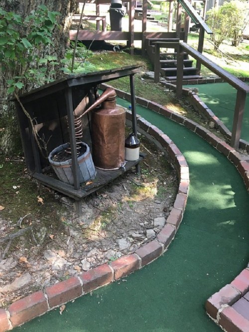 A rustic display with a copper still, barrel, and bottle beside a winding mini-golf path surrounded by greenery.
