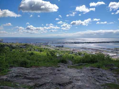 A scenic view of a coastal landscape with green hills, a blue sky, and clouds over a shimmering body of water.