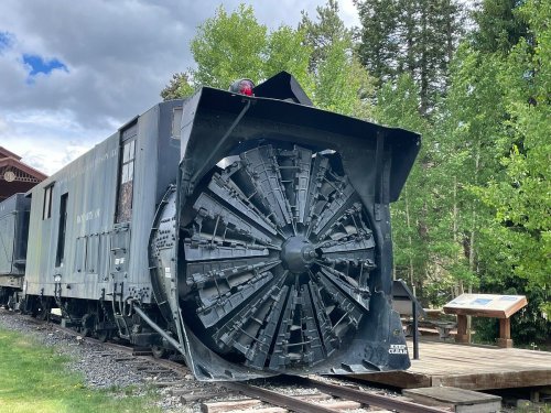 A snowplow train car with a large rotating blade, parked near a wooden platform surrounded by trees.