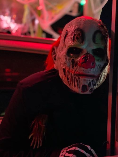 A person wearing a creepy clown mask with red hair, sitting in low light with eerie decorations in the background.