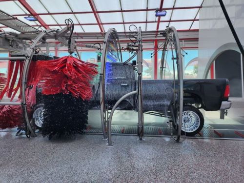 A black truck in a car wash, surrounded by colorful brushes and water spray, with a modern facility in the background.