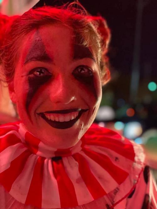 A smiling clown with red and white makeup, wearing a striped collar, illuminated by red light.