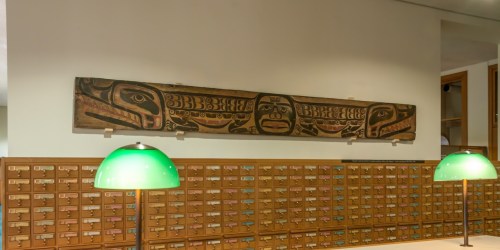 A wooden totem with carved figures hangs above a card catalog with green lamps in a library setting.
