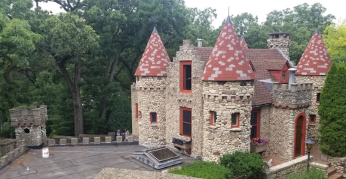 A stone castle with red-tiled roofs and turrets, surrounded by trees and greenery.