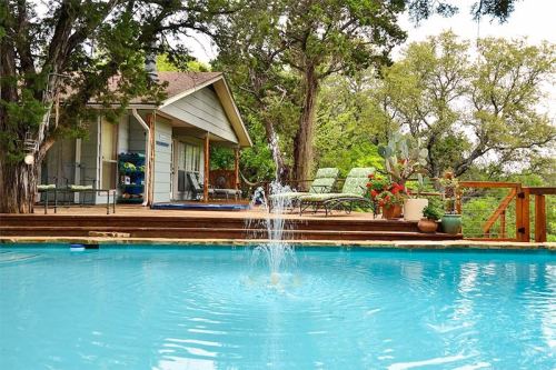 A serene backyard with a pool, fountain, and a cozy house surrounded by trees and potted plants.