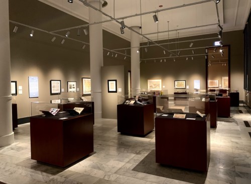 A spacious art gallery with wooden display cases showcasing various artworks and framed pieces on the walls.