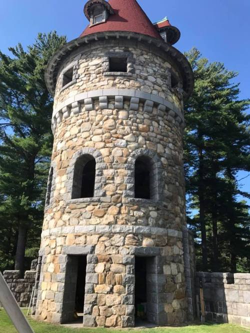 A tall stone tower with a red roof, surrounded by trees under a clear blue sky.