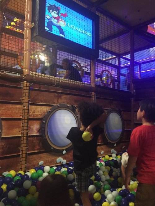 Children playing in a colorful ball pit with a screen displaying "LOADING" above them in a fun indoor play area.