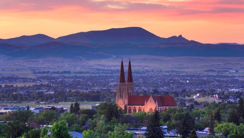 A scenic view of a church with spires at sunset, surrounded by mountains and a valley landscape.