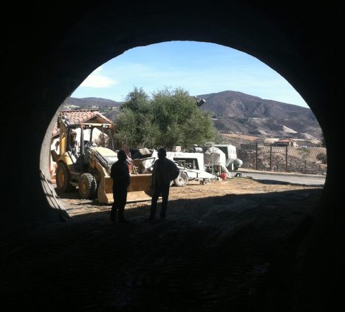 Two workers stand in a tunnel entrance, observing construction equipment and a scenic landscape beyond.