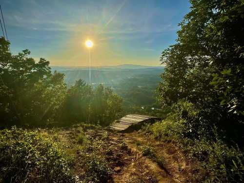 A sunlit landscape from a mountain trail, with lush greenery and a view of distant hills under a clear blue sky.