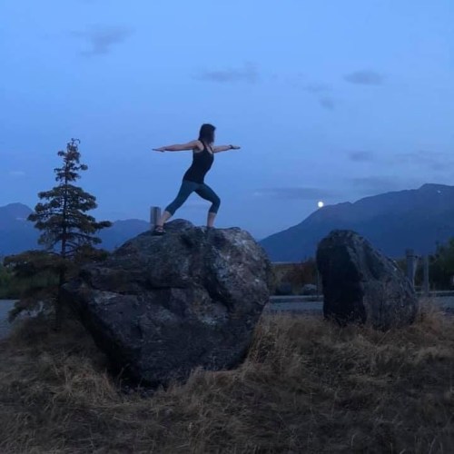 A person practicing yoga on a rock at dusk, with mountains and a rising moon in the background.