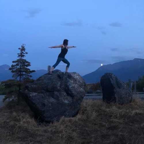 A person practicing yoga on a rock at dusk, with mountains and a rising moon in the background.