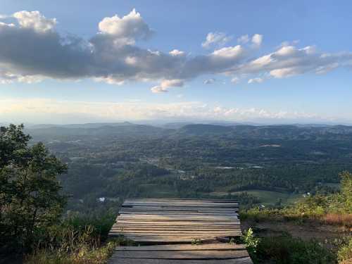 A wooden platform overlooks a vast valley with rolling hills and a cloudy sky in the background.