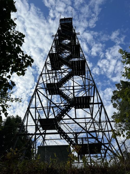 A tall metal observation tower against a blue sky with scattered clouds, surrounded by trees and a fence.