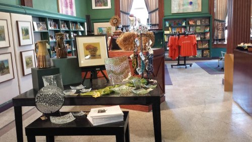 A cozy shop interior featuring art, crafts, and books, with a display table of decorative glass and textiles.