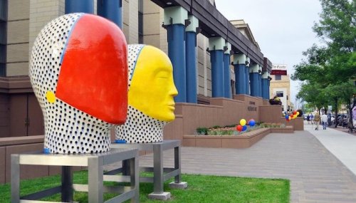 Colorful abstract sculptures of heads in red and yellow, placed outside a modern building with blue columns.