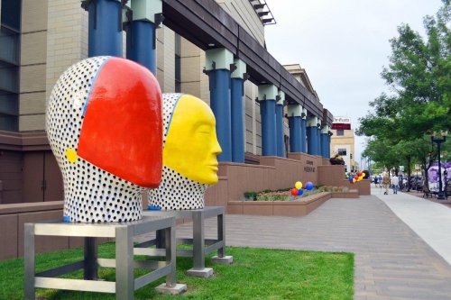 Colorful abstract sculptures of heads in red and yellow, set against a modern building with blue columns and greenery.